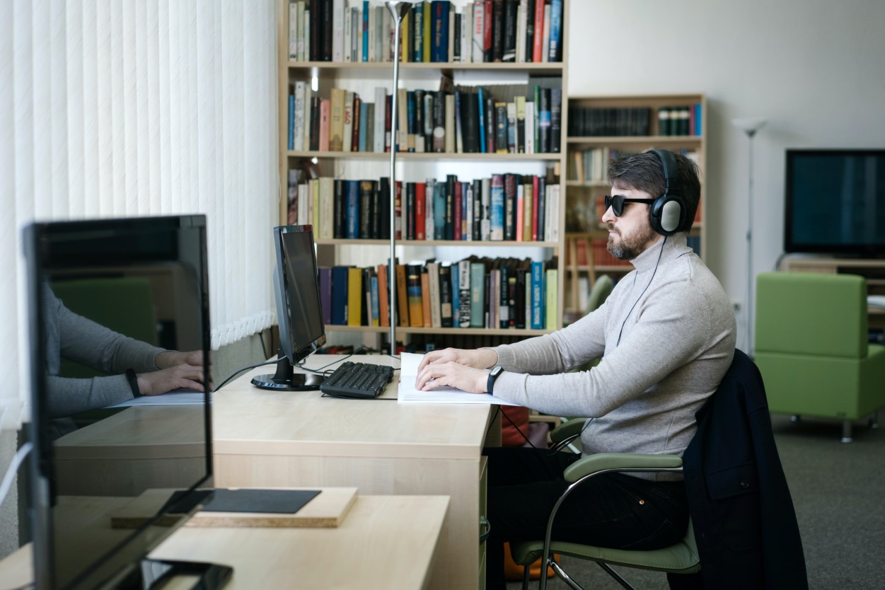 A man who is blind or low sighted wearing a sweater and headphones, sitting in front of a computer in a library, reading a braille book.